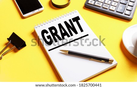 The word GRANT is written in a white notebook on a yellow background near a pen, phone, coffee and other office supplies.