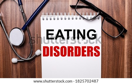 EATING DISORDERS is written on white paper on a wooden background near the stethoscope and black-framed glasses. Medical concept