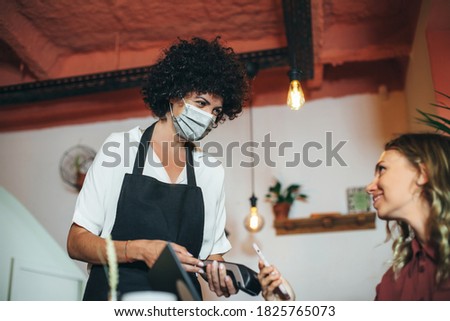 Mobile payment with wallet app and wireless nfc technology. Smiling woman paying with smartphone application and credit card information. Waitress smiling with protective face mask during coronavirus.