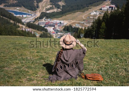 woman in a poncho, hat and with a backpack, back to the camera, looking at the mountains