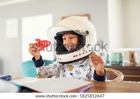 Little boy wearing an astronaut helmet costume and playing with a spaceship while doing homework. Cute kid in astronaut pajamas with toy rocket playing and dreaming of becoming a spacemen. Royalty-Free Stock Photo #1825812647