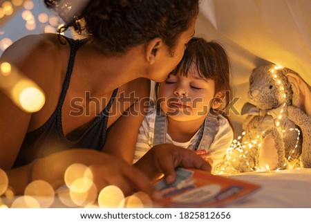 African american mother kissing her daughter on forehead while lying on bed in illuminated tent. Mother putting daughter to sleep in cozy hut after reading a fairy tale in tent. Mom kissing cute girl.