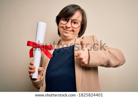 Young down syndrome business woman holding univeristy diploma award over isolated background happy with big smile doing ok sign, thumb up with fingers, excellent sign