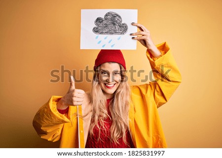Young beautiful blonde woman wearing raincoat and wool cap holding banner with cloud image happy with big smile doing ok sign, thumb up with fingers, excellent sign
