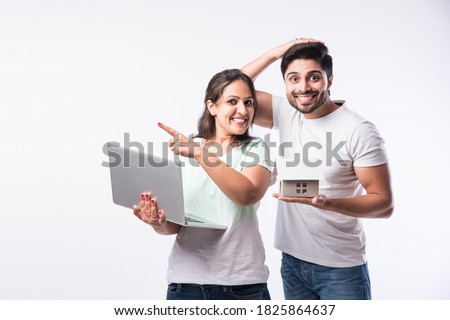 Indian young family couple choosing new home online, search real estate to buy or rent, house for sale on screen of computer, standing against white background
