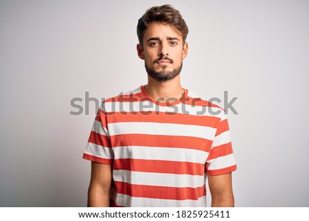 Young handsome man with beard wearing striped t-shirt standing over white background Relaxed with serious expression on face. Simple and natural looking at the camera. Royalty-Free Stock Photo #1825925411