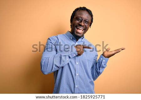 Young handsome african american man wearing shirt and glasses over yellow background amazed and smiling to the camera while presenting with hand and pointing with finger.