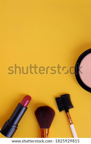 Makeup brush and cosmetics on yellow background with copyspace. Pink lipstick, compact powder, brush and brow comb. Vertical photo.  