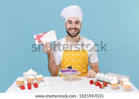 Smiling young bearded male chef or cook baker man in apron white t-shirt toque chefs hat cooking at table isolated on blue background. Cooking food concept. Mock up copy space. Hold gift certificate
