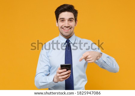 Smiling young business man in classic blue shirt tie posing isolated on yellow background studio. Achievement career wealth business concept. Mock up copy space. Pointing index finger on mobile phone