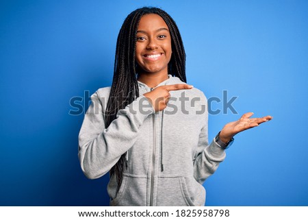 Young african american athlete woman wearing sports sweatshirt over blue isolated background amazed and smiling to the camera while presenting with hand and pointing with finger.