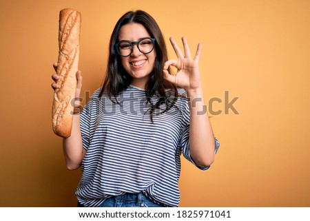 Young brunette woman holding homemade fresh bread baguette over yellow background doing ok sign with fingers, excellent symbol