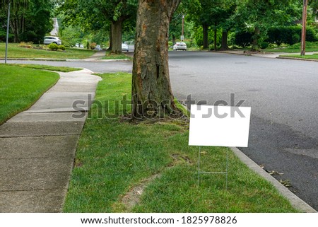 A white blank advertisement sign on a grassy strip near a street and a tree trunk