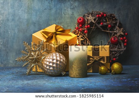 Beautiful composition of golden gift box with shiny satin bow for present, candle, wreath, toys for christmas eve on blue concrete background