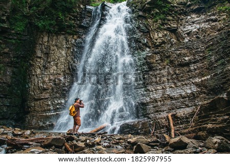 Traveler man with a yellow backpack standing on the background of a waterfall makes a photo landscape. Travel lifestyle concept. Copy, empty space for text