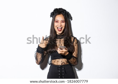 Portrait of satisfied asian woman in elegant gothic dress and black wreath showing thumbs-up while using mobile phone, standing over white background