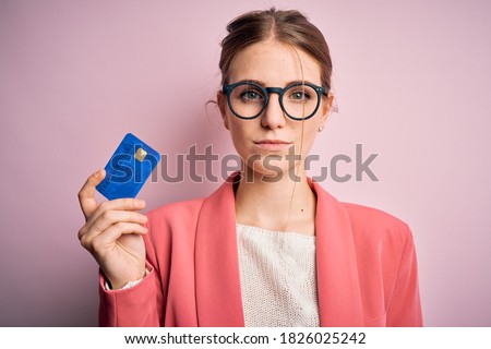 Young beautiful redhead woman holding credit card over isolated pink background with a confident expression on smart face thinking serious
