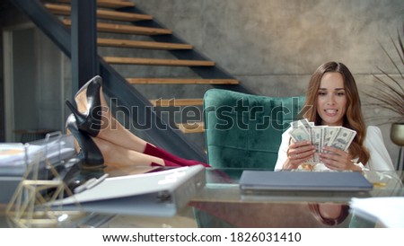 Successful woman celebrating good deal at desk in slow motion. Portrait of business woman making money rain in office. Happy woman throwing money away in workplace. Girl putting legs on table.