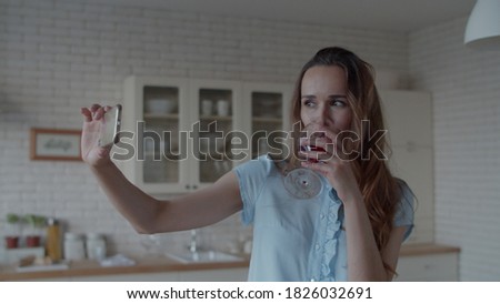 Close up young woman posing for photo with glass of red wine in kitchen interior in slow motion. Happy woman drinking wine in front of phone camera in kitchen. 