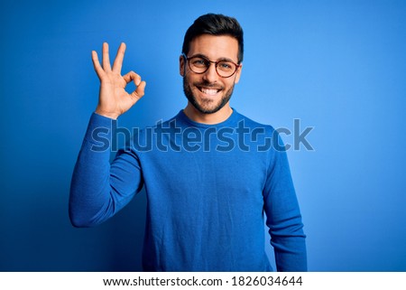 Young handsome man with beard wearing casual sweater and glasses over blue background smiling positive doing ok sign with hand and fingers. Successful expression.