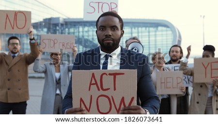 Portrait of African American man in suit and tie standing outdoor at protest with poster Act Now. Male protester at strike against unemployment. Mixed-races people on background. Protesting. Striking.