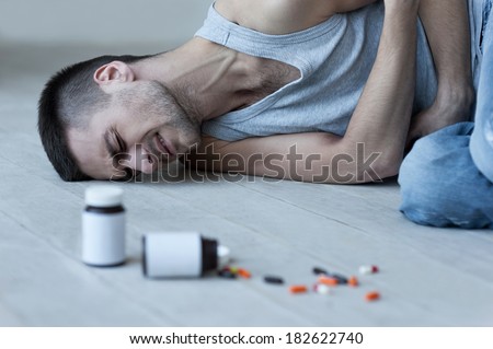 Numbing his pain. Young man lying on the floor and holding hands on stomach while bottles with pills laying near him