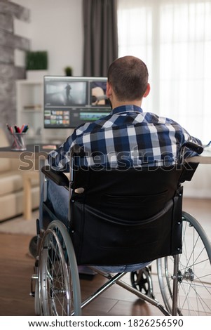 Disabled video editor sitting on wheelchair doing post production on film. Invalid videographer doing his business online, using high technology, sitting in his apartment, working remotely in special
