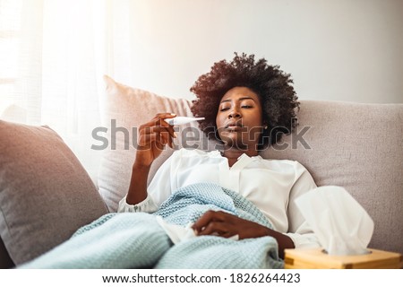 Woman with flu in bed, she use home medicine to handle sickness. Sickness, seasonal virus problem concept. Woman being sick having flu lying on sofa looking at temperature on thermometer.  Royalty-Free Stock Photo #1826264423
