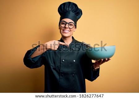 Young beautiful brunette chef woman wearing cooker uniform and hat holding bowl and whisk very happy pointing with hand and finger