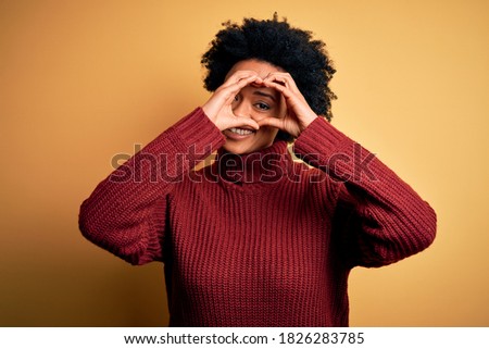 Young beautiful African American afro woman with curly hair wearing casual turtleneck sweater Doing heart shape with hand and fingers smiling looking through sign