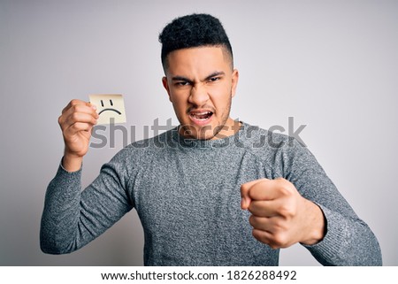 Handsome man holding reminder paper with sad emotion face emoji over white background annoyed and frustrated shouting with anger, crazy and yelling with raised hand, anger concept