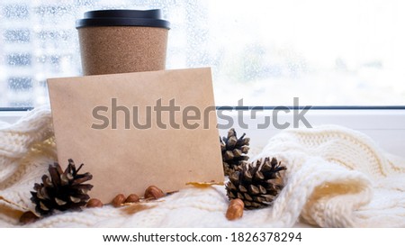 A cup for coffee near the window, a knitted scarf, pine cones, a letter. Autumn and winter background.