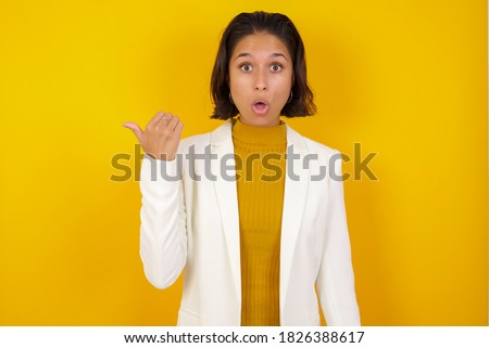 Horizontal shot of pretty young joyful shocked businesswoman points with thumb away, indicates something on blank wall isolated over white background. Check this out. Ethnicity concept.