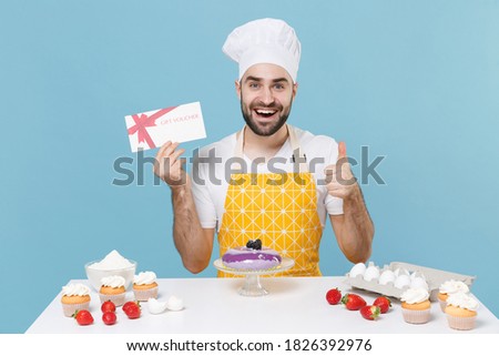 Excited young bearded male chef or cook baker man in apron white t-shirt toque chefs hat cooking at table isolated on blue background. Cooking food concept. Hold gift certificate, showing thumb up