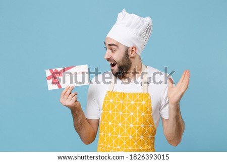 Excited young bearded male chef or cook baker man in apron white t-shirt toque chefs hat isolated on blue background. Cooking food concept. Mock up copy space. Hold gift certificate spreading hands