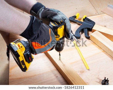 Man hands in safety gloves is screwing the screw with the cordless drill. Instruments and wooden details around. Close up.  Royalty-Free Stock Photo #1826412311