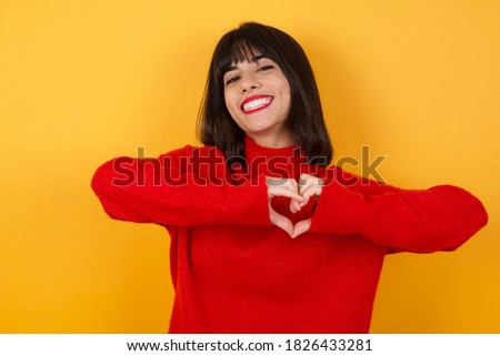 Caucasian brunette woman wearing red casual sweater isolated over yellow background smiling in love doing heart symbol shape with hands. Romantic concept.