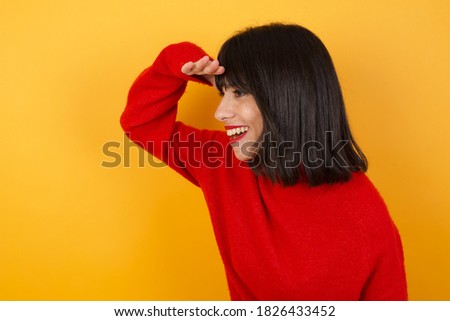 Caucasian brunette woman wearing red casual sweater isolated over yellow background very happy and smiling looking far away with hand over head. Searching concept.