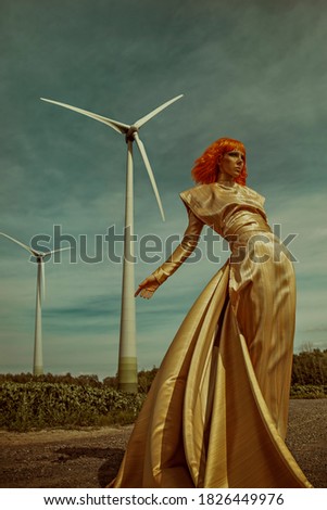 Young woman with an orange bob haircut, wearing a long shiny golden dress, standing in front of a corn field and wind turbines on a sunny day
