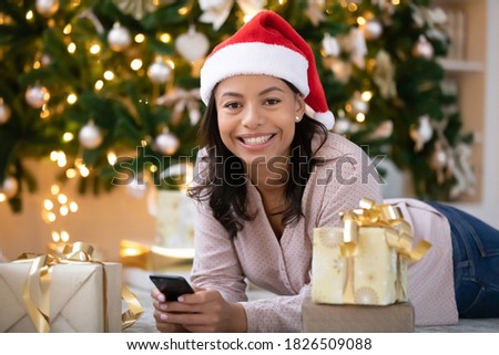 Portrait of smiling african American woman in santa hat lying under Christmas tree with wrapped gifts using smartphone, happy biracial female text holiday new year greeting on cellphone gadget