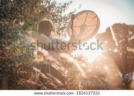 A traditional slavic shaman girl beating the drum, evoking the spirits of nature in a lovely sunset field scenery Royalty-Free Stock Photo #1826537222