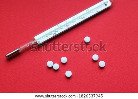 A mercury glass thermometer and seven small round tablets lie on a bright red surface. The thermometer lies with numbers up. Treatment. Medicines.