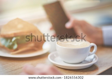 Energy drink. Cropped shot of a man using smartphone while having breakfast in cafe. Cup of coffee and fresh sandwich on wooden table