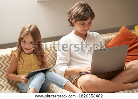 Happy little boy and girl, brother and sister sitting on sofa at home, using laptop and digital tablet and watching cartoons or playing games online