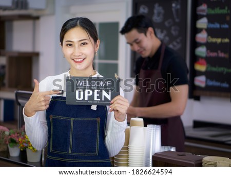 Young Asian cafe owner pointed at the opening sign held in hand with a smile. The barista prepares the equipment in the back. Morning atmosphere in a coffee shop.