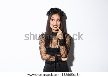 Image of intrigued young cute woman in witch dress, celebrating halloween, thinking about something interesting, looking upper left corner, standing over white background