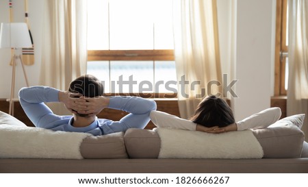 Finding zen. Rear back view of young family male and female newlyweds resting on cozy sofa in suite room of luxurious hotel listening to quiet calm music and admiring beautiful scenery out of window Royalty-Free Stock Photo #1826666267