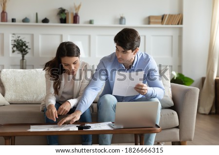 Payday. Concentrated attentive millennial spouses sitting on couch at home discussing possibility of taking out loan or credit, counting sum of taxes or bills to pay online using laptop and calculator Royalty-Free Stock Photo #1826666315