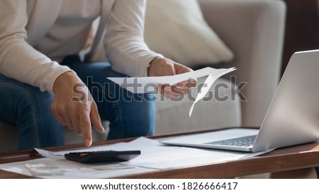 Home office. Close up of young woman owner or renter of dwelling sitting on sofa at desk before laptop screen making calculations of utility payment holding paper bill, invoice or notification in hand Royalty-Free Stock Photo #1826666417
