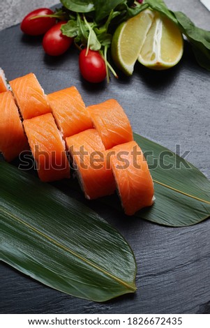 A set of sushi rolls lies on bamboo leaves with lime, herbs and red tomatoes, served on a black stone slate. Dark background